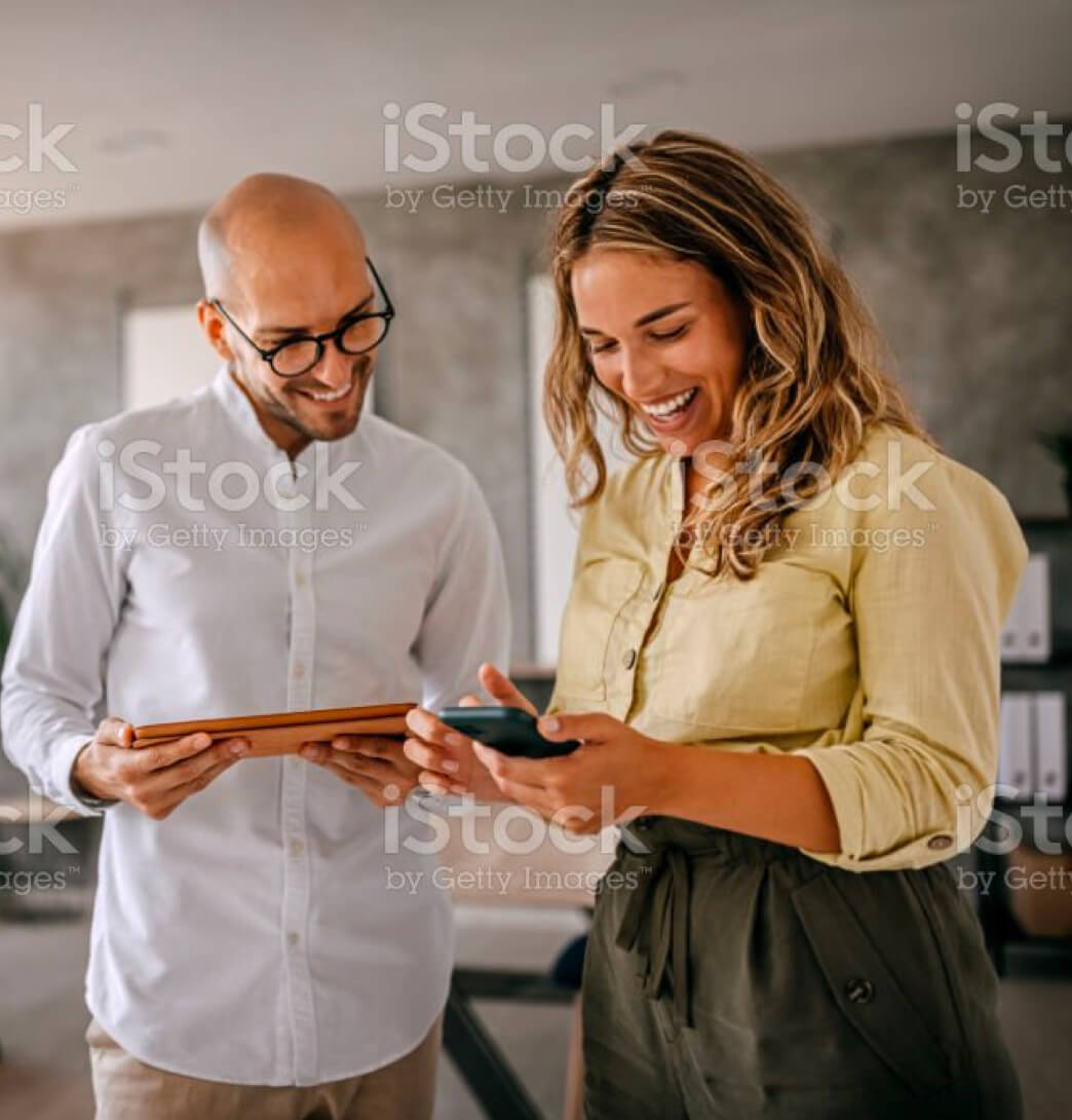 Cheerful and smiling young successful female businesswoman standing with colleague looking at smartphone in modern office and coworking space